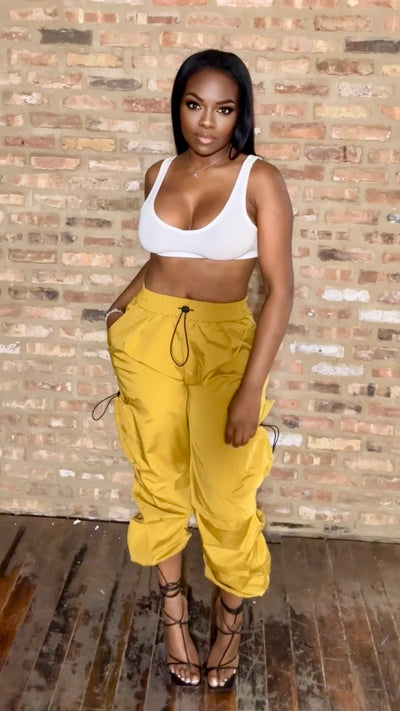 Woman wearing a white crop top and yellow pants standing against a brick wall.